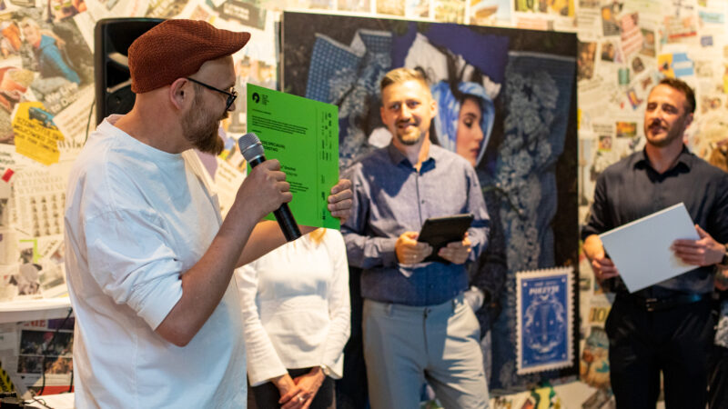 Na pierwszym planie widać jednego z laureatów konkursu - mężczyznę w białej koszulce i brązowym kaszkiecie. Mężczyzna trzyma dyplom. W głębi widać kilku jurorów konkursu (2 mężczyzn oraz kobietę).